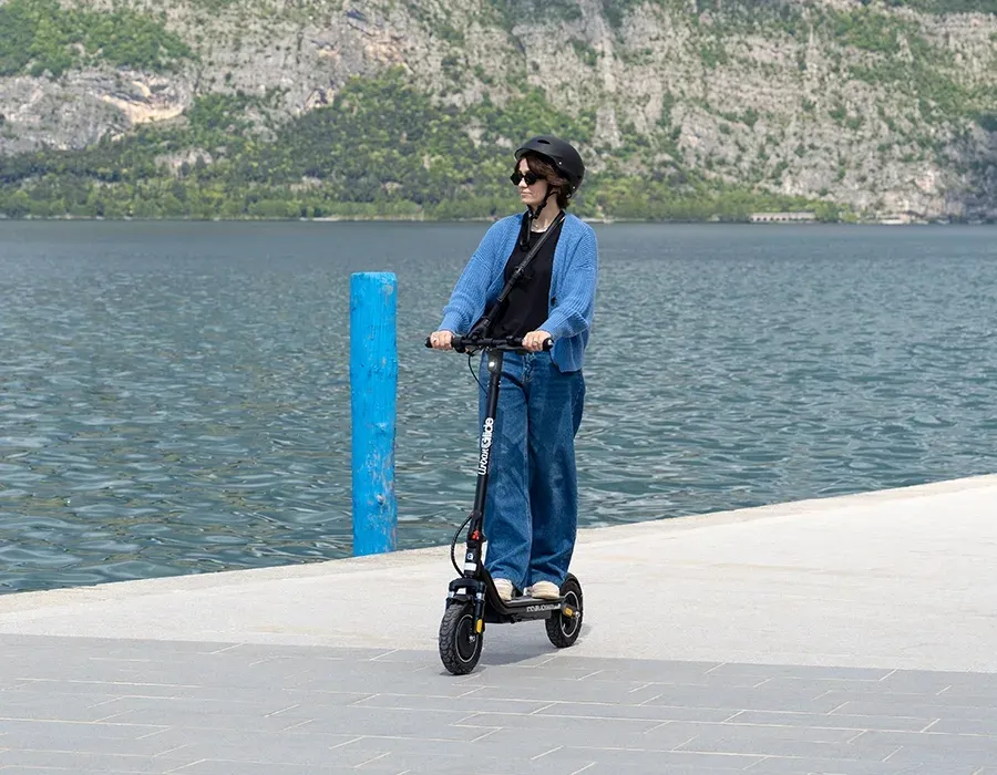Une femme casquée roule en Urbanglide 100 Evo sur un quai dallé au bord d'un lac